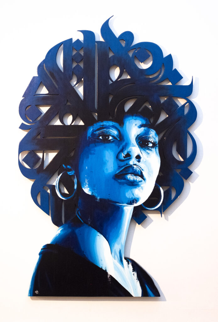 Portrait de femme noire en différentes teintes de bleu. Le portrait est découpé dans du bois et les cheveux sont formés par de la calligraphie